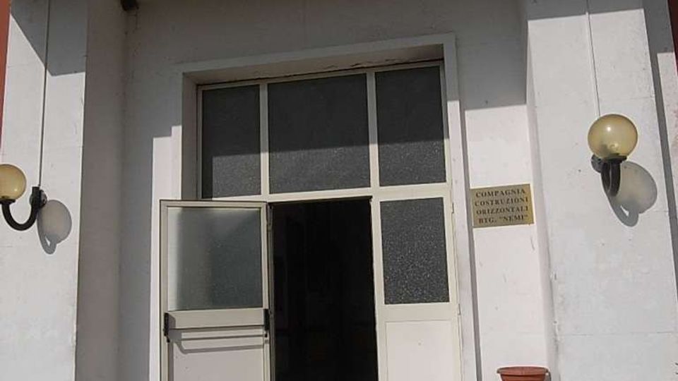 Ingresso di un edificio con facciata bianca e portico sporgente, raggiungibile tramite alcuni gradini. Porta a due ante con vetri opachi e sopraluce, lampade a parete ai lati e una piccola targa sulla destra.