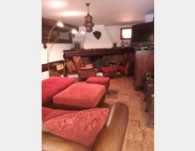 Salotto rustico con pavimento in piastrelle, grande camino in muratura con trave in legno e decorazioni, e lampadario sospeso in stile marocchino. Divani e poltrone in pelle con cuscini rossi e pouf sono disposti attorno al camino, con un mobile in legno e un televisore a lato.
