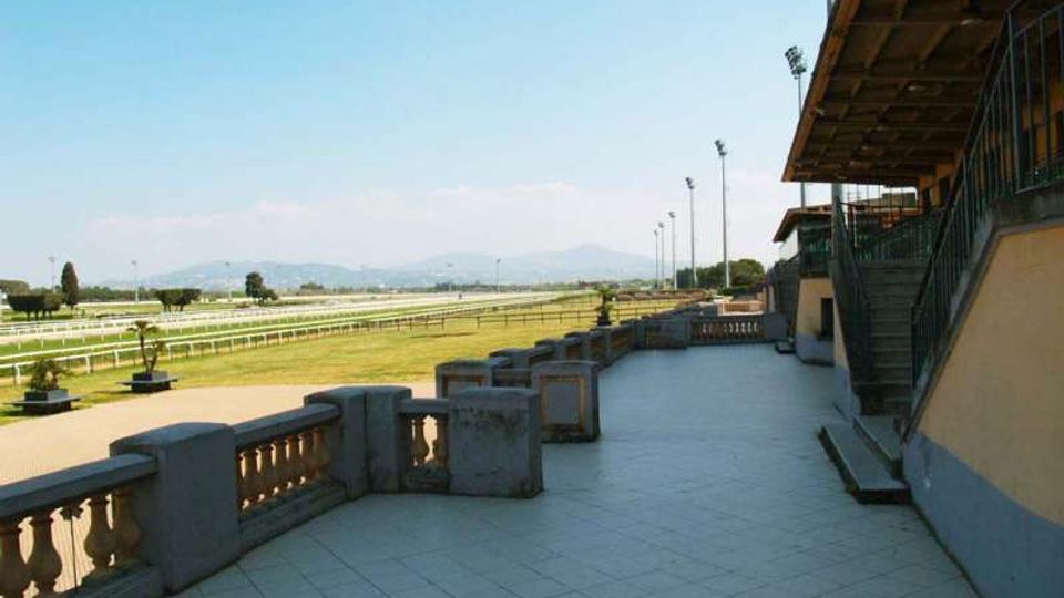 Terrazza pavimentata con balaustra in pietra accanto a una tribuna coperta con scale esterne e ringhiere metalliche. Sullo sfondo si vedono i rettilinei di una pista ippica con prato, pali dell’illuminazione e colline lontane sotto cielo sereno.