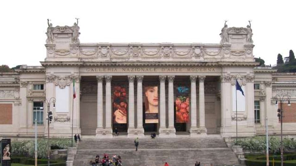 Facciata monumentale in stile neoclassico della Galleria Nazionale d’Arte Moderna, con ampio portico a colonne, grandi scalinate centrali e banner verticali appesi tra le colonne. Davanti si vede un’area verde con vialetti e lampioni, con bandiere ai lati dell’ingresso.