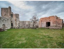 Cortile erboso all’interno di un complesso di rovine in pietra, con mura alte e aperture ad arco tipiche di una struttura fortificata o castello. Sullo sfondo si vedono un albero spoglio, recinzioni basse e un cielo nuvoloso.