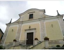 Facciata di una chiesa in stile semplice con intonaco giallo e cornici in pietra chiara, timpano curvilineo e una finestra centrale superiore. In basso si vede l’ingresso con portale scuro, una scalinata con ringhiera in ferro e vasi di piante su pilastri, sotto un cielo nuvoloso.