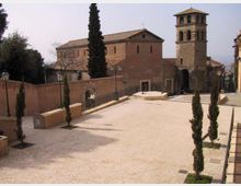 Piazza lastricata in pietra con aiuole e cipressi, circondata da muretti e lampioni. Sullo sfondo si vede una chiesa in mattoni con campanile in pietra e tetti in coppi, affacciata su un leggero pendio urbano.