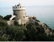 Torre fortificata in pietra con corona merlata e piccola cupola bianca, collegata a mura difensive su un promontorio roccioso. Sullo sfondo si vede il mare aperto, con vegetazione mediterranea in primo piano.