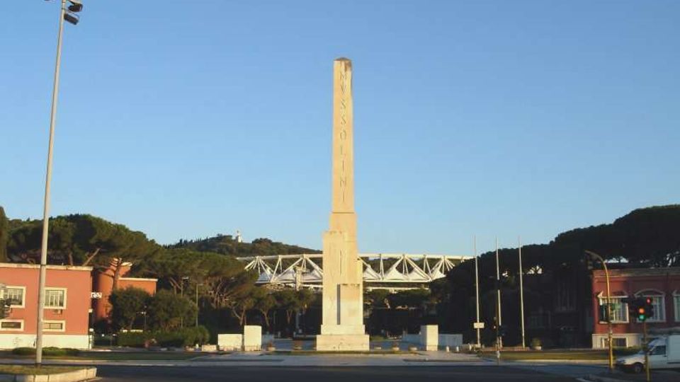 Rotonda stradale con un alto obelisco in pietra al centro, circondata da carreggiate e pali dell’illuminazione. Sullo sfondo si vedono edifici bassi, alberi e una struttura metallica simile a un ponte sotto un cielo sereno.