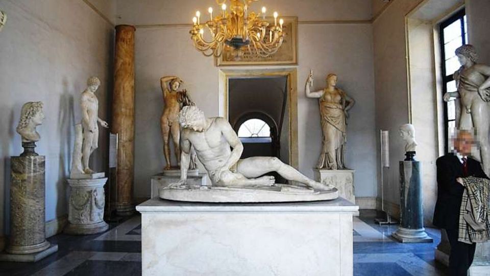 Sala museale con pavimento a scacchi e lampadario dorato, con al centro un grande basamento che sostiene una statua in marmo distesa. Lungo le pareti sono esposte diverse sculture classiche su piedistalli, con finestre alte e un passaggio ad arco sullo sfondo.
