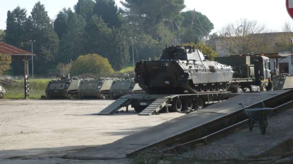 Piazzale asfaltato in un’area militare con un carro armato su un rimorchio a pianale ribassato collegato a una rampa metallica. Sullo sfondo si vedono altri mezzi corazzati in fila, edifici bassi e alberi lungo il perimetro.