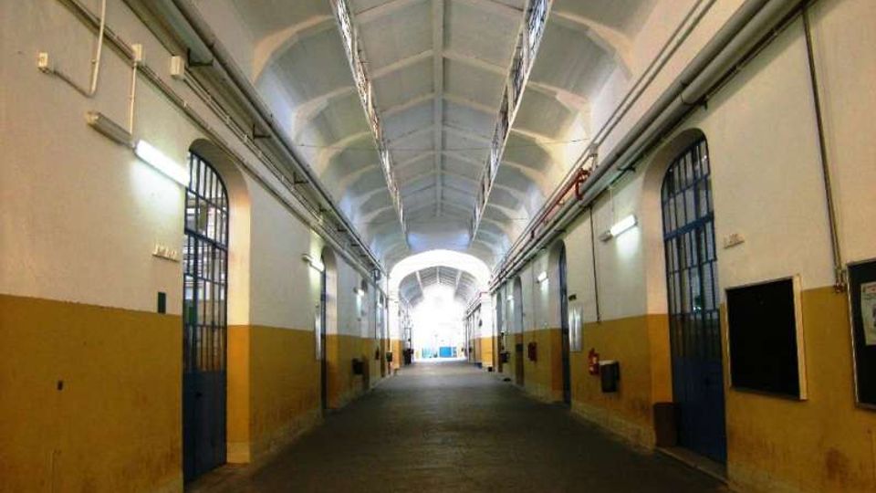 Lungo corridoio interno con soffitto a volta e archi ripetuti, pareti bicolori bianco e ocra e tubazioni a vista lungo i lati. Ai lati si trovano porte metalliche con grate e lampade a parete, con un’uscita luminosa in fondo.