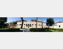 Ampio edificio storico color crema con persiane verdi e frontone decorato centrale, affacciato su un giardino con siepi potate, alberi alti e una vasca in pietra in primo piano. Sullo sfondo si vedono altri corpi di fabbrica e una torre in muratura, in un contesto di parco.