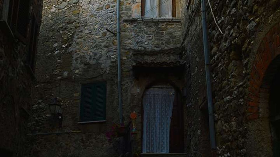 Vicolo stretto tra edifici in pietra con scala in muratura che sale verso un portone ad arco con tenda. Facciate rustiche con persiane e finestre ad arco, grondaie a vista e tetto con tegole.