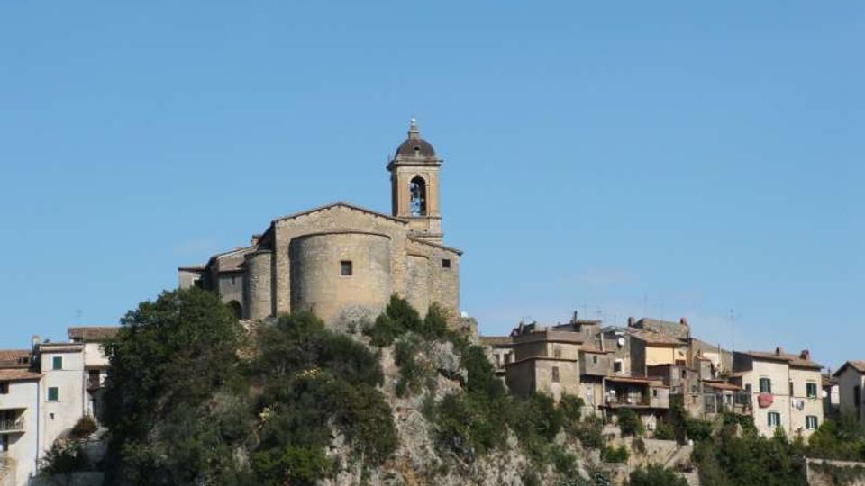 Chiesa in pietra con campanile su una sommità rocciosa, affiancata da un borgo di case addossate alla scogliera. Vegetazione arbustiva sul pendio e cielo sereno sullo sfondo.