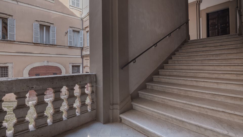 Scalinata interna in pietra chiara con corrimano in ferro che sale verso un portone scuro sotto archi, affacciata su un cortile tramite una balaustra in muratura. Sullo sfondo si vedono facciate di edificio con finestre e persiane azzurre.