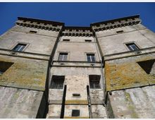 Vista dal basso della facciata di un castello in pietra con due torri laterali e corpo centrale, finestre rettangolari e merli sotto la gronda. Le pareti mostrano intonaco e pietra invecchiati con tracce di muschio, sullo sfondo di un cielo azzurro.