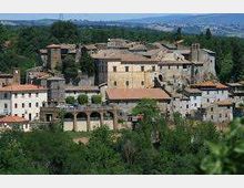 Veduta panoramica di un borgo storico su una collina, con edifici in pietra dai tetti in tegole e una struttura ad archi nella parte inferiore. Il paese è circondato da colline verdi e vegetazione, con l’orizzonte della campagna sullo sfondo.