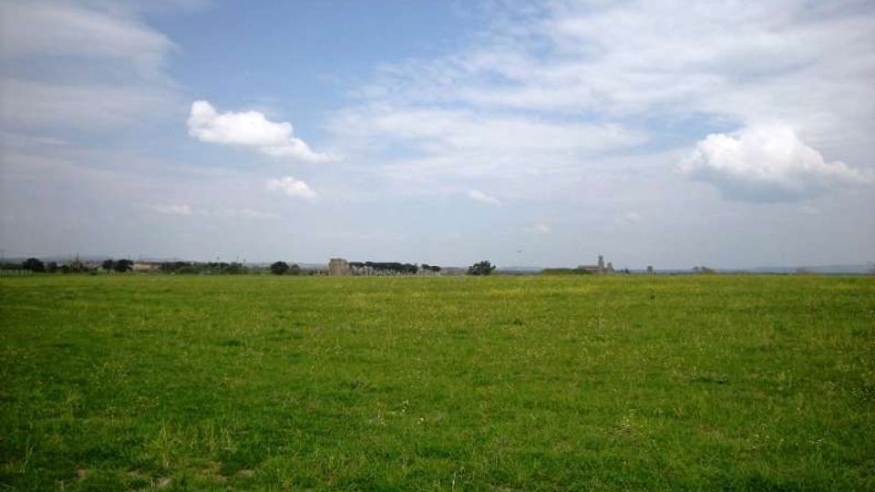 Ampia pianura erbosa con prato verde che si estende fino all’orizzonte, con pochi edifici lontani e alberi sparsi. Sopra, un cielo aperto con nuvole bianche e sottili velature.