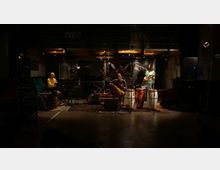 Interno di una sala prove o piccolo studio musicale con palco basso e pavimento lucido, illuminato da luci calde a soffitto. Al centro si vedono batteria, tastiera e strumenti a percussione con microfoni e cavi, affiancati da grandi casse acustiche ai lati.