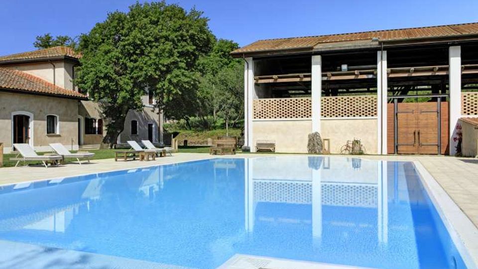 Piscina rettangolare all’aperto con acqua azzurra e bordo in pietra, circondata da lettini e prato. Sullo sfondo si vedono edifici rustici con tetti in coppi e un grande portico aperto con colonne bianche, in un contesto di campagna con alberi.