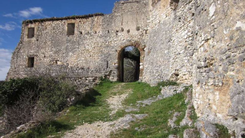 Mura in pietra di una fortificazione in rovina con un grande arco d’ingresso e piccole aperture, affacciate su un sentiero sterrato con tratti d’erba. La scena è all’aperto, su un pendio roccioso, sotto un cielo azzurro.