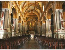 Interno di una chiesa barocca con navata centrale fiancheggiata da file di panche in legno, colonne e archi decorati. La volta e le pareti sono riccamente ornate con dorature e affreschi, con l’altare maggiore in fondo illuminato.
