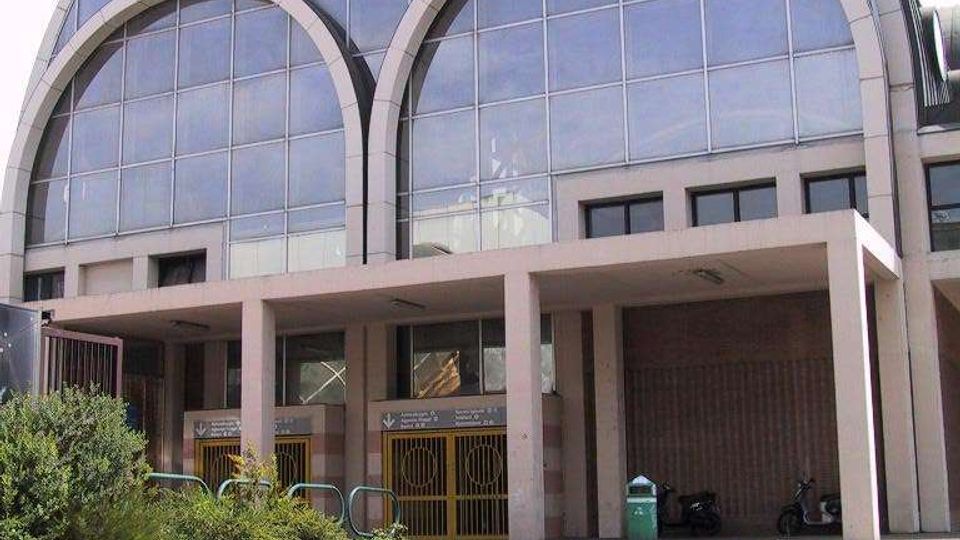 Facciata di un grande edificio moderno con ampie vetrate a griglia e copertura ad arco, caratterizzata da due grandi archi decorativi sovrapposti. Al piano terra si vedono un portico con pilastri, ingressi con cancelli e un piazzale asfaltato con aiuole.