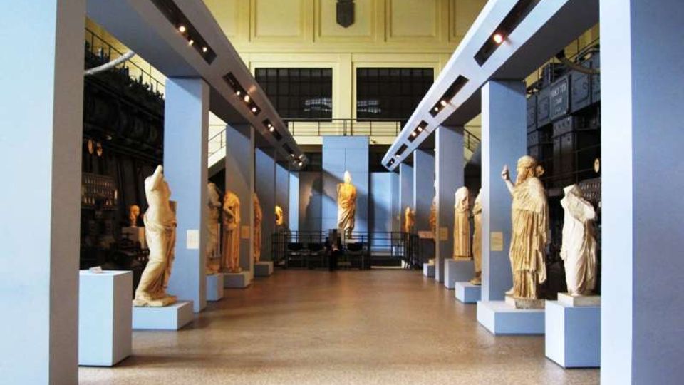 Interno di una sala museale con lungo corridoio centrale, strutture espositive azzurre e statue classiche su piedistalli ai lati. In fondo si vede una grande parete con finestre alte e illuminazione a soffitto.
