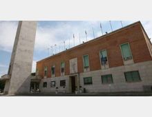 Facciata di un edificio istituzionale in mattoni rossi con basamento in pietra chiara, finestre rettangolari e ingresso centrale, con una fila di pennoni sul tetto. In primo piano si vede un alto obelisco in pietra e un’area pavimentata davanti all’edificio.