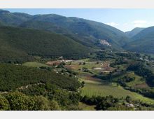 Veduta panoramica di una valle montana con campi coltivati e filari regolari, strade sinuose e piccoli nuclei di edifici sparsi. Sullo sfondo si vedono ampi versanti boscosi che circondano la pianura agricola.