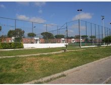 Campo sportivo all’aperto recintato con alte reti metalliche e pali di illuminazione, circondato da un prato e un vialetto asfaltato. Sullo sfondo si vedono alberi e edifici bassi con cielo parzialmente nuvoloso.
