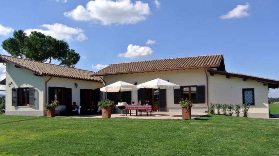 Casa rurale a un piano con tetto in coppi, persiane scure e veranda centrale con due ombrelloni e tavolo. Davanti si estende un ampio prato verde con vasi di piante e alberi sullo sfondo sotto un cielo azzurro con nuvole.