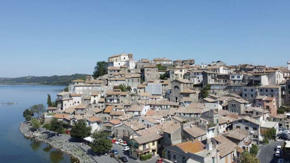 Vista panoramica di un borgo collinare con case in pietra dai tetti in coppi, addossate l’una all’altra, e una chiesa in posizione elevata. Il paese si affaccia su un lago con riva alberata e strada lungo l’acqua sotto un cielo sereno.