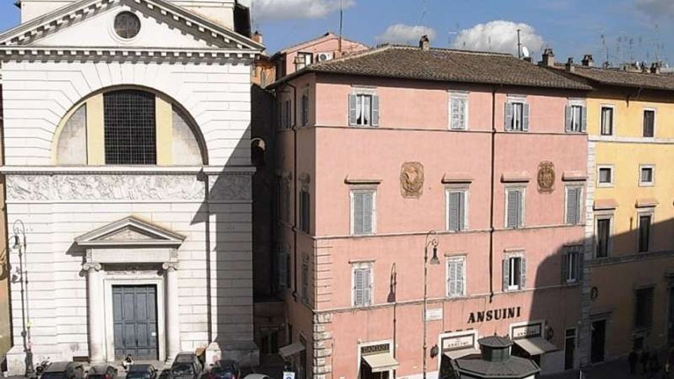 Facciate su una piazza urbana: a sinistra un edificio bianco in stile neoclassico con grande frontone e portale, a destra un palazzo rosa a più piani con persiane e insegna “ANSUINI”. In basso si vedono auto parcheggiate lungo il bordo della piazza e lampioni stradali.