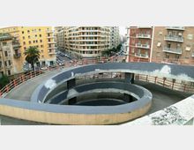 Rampa pedonale sopraelevata a spirale in cemento, con parapetti metallici, vista dall’alto. Sullo sfondo si vedono strade urbane e palazzi residenziali multipiano con balconi.