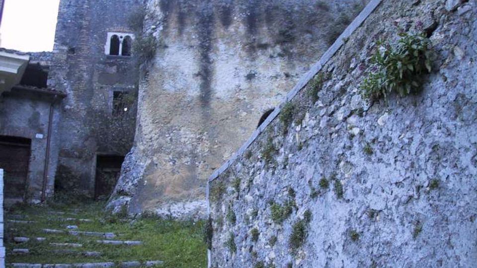 Cortile interno in pietra con gradini irregolari che salgono tra muri alti e scrostati, con tracce di umidità e vegetazione spontanea. Sullo sfondo si vede un edificio in muratura con piccole finestre ad arco e un passaggio scuro.