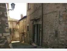 Vicolo stretto in discesa con gradini in pietra tra edifici in muratura di pietra e mattoni, con porte e finestre dai telai scuri. Un lampione a parete e cavi esterni corrono lungo le facciate, in un contesto di borgo storico.