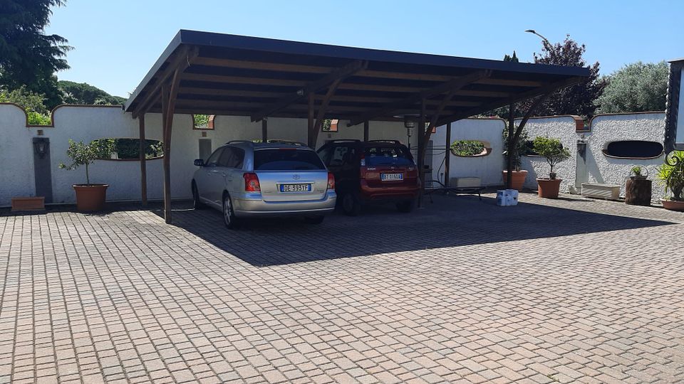 Parcheggio pavimentato in autobloccanti con una tettoia in legno a falda unica che copre due posti auto. Sullo sfondo un muro bianco perimetrale con nicchie e vasi di piante, circondato da alberi sotto cielo sereno.