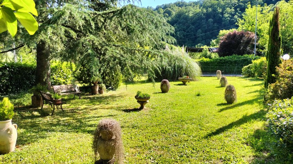 Giardino ampio con prato curato, grandi alberi ombrosi e panchine, con vasi e piccoli elementi decorativi disposti lungo un vialetto erboso. Sullo sfondo si vedono siepi e un pendio boscoso sotto cielo sereno.