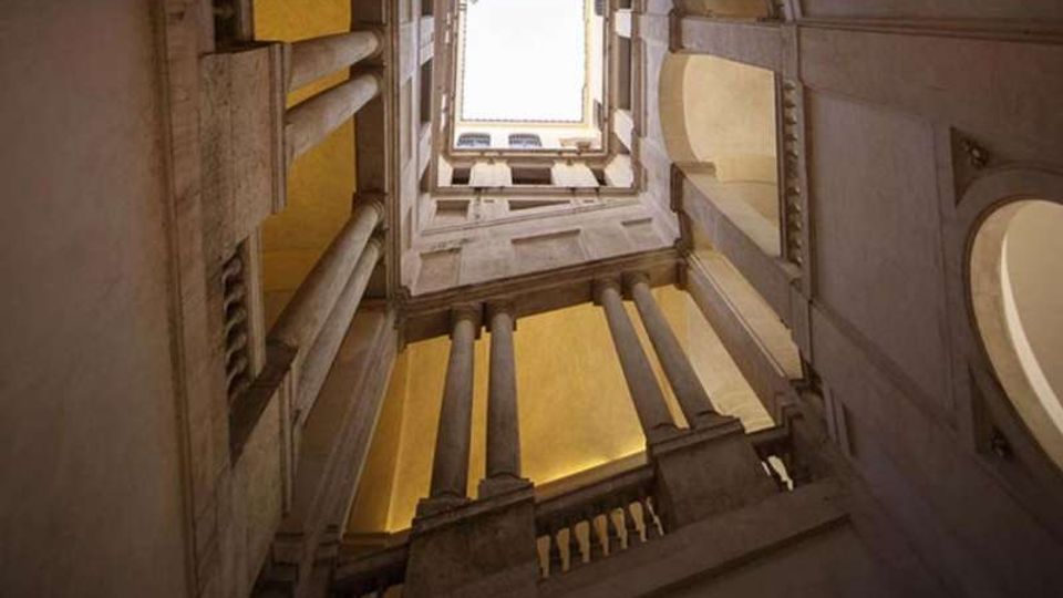 Veduta dal basso all’interno di una corte verticale in pietra, con colonne e balaustre su più livelli, che conduce a un lucernario quadrato aperto verso il cielo. Le pareti presentano archi e aperture laterali illuminate da luce calda.