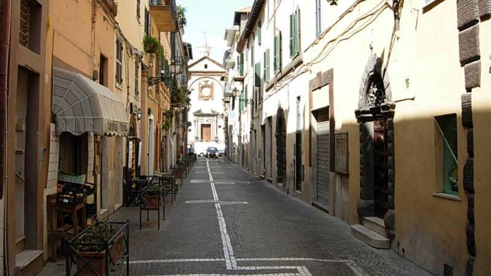 Strada stretta lastricata in un centro storico, fiancheggiata da edifici alti con persiane e balconi, con tavolini all’esterno e cavi/lanterne lungo le facciate. In fondo alla via si vede la facciata di una chiesa con portale centrale e decorazioni.