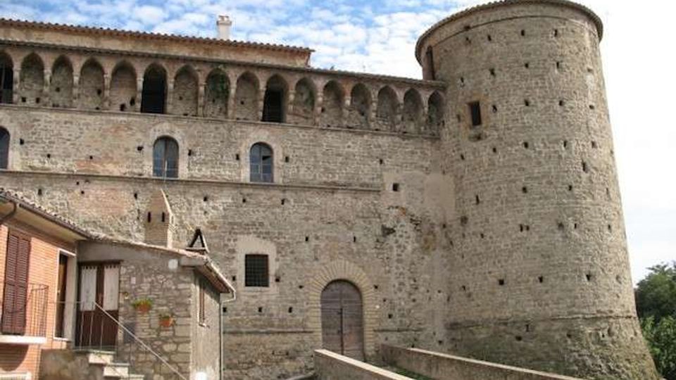 Facciata in pietra di un castello medievale con torre cilindrica merlata e loggiato ad arcate lungo il livello superiore. In primo piano si vedono una rampa murata e una piccola costruzione addossata alle mura, sotto un cielo parzialmente nuvoloso.