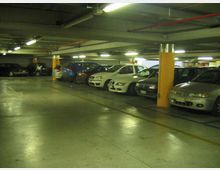 Parcheggio sotterraneo con file di auto parcheggiate lungo le pareti e una corsia centrale libera. Soffitto basso con tubazioni a vista e illuminazione al neon, pilastri con fasce gialle e pavimento in cemento con strisce di delimitazione.