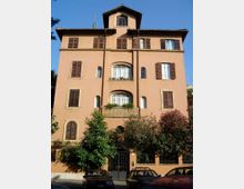 Facciata di un palazzo residenziale a più piani intonacato color ocra, con numerose finestre dotate di persiane in legno e balconi ad arco centrali. In primo piano si vedono un cancello d’ingresso con recinzione, alberi e cespugli lungo la strada.