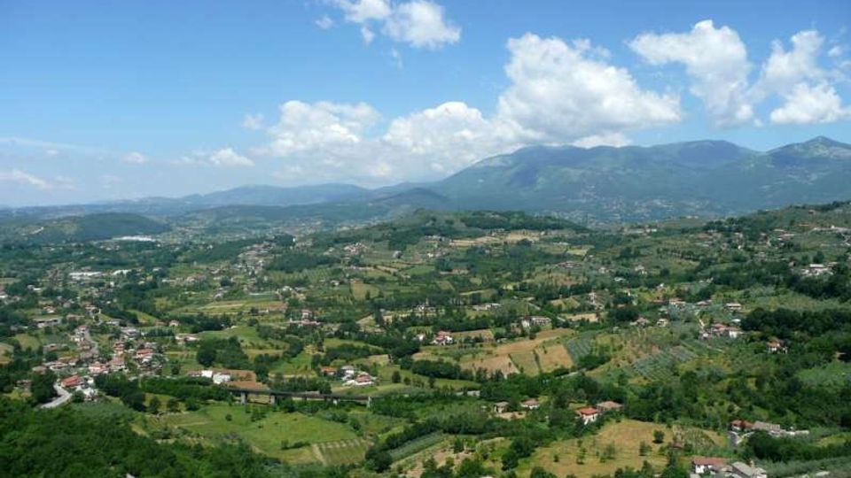 Veduta panoramica di una valle collinare con campi coltivati, boschi e piccoli borghi sparsi. Sullo sfondo si vedono montagne verdi sotto un cielo azzurro con nuvole.