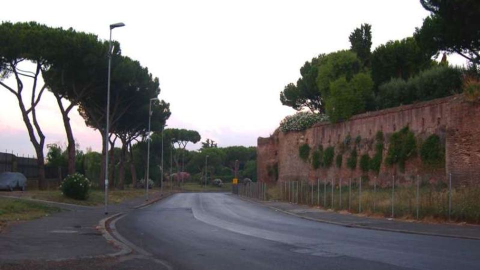 Strada asfaltata in lieve curva con marciapiede e lampioni, fiancheggiata da alti pini mediterranei. Sulla destra corre un lungo muro in mattoni coperto in parte da vegetazione, con recinzione alla base.