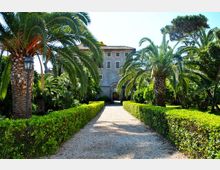 Viale in ghiaia fiancheggiato da siepi potate e alte palme in un giardino mediterraneo, che conduce a una villa storica in pietra con tetto in tegole e arco d’ingresso centrale. Vegetazione rigogliosa e lampioni classici lungo il percorso.