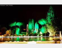 Fontana rettangolare illuminata di notte, con getti d’acqua lungo il bordo e una vasca centrale. Sullo sfondo un giardino con alberi e siepi alti illuminati da luci verdi e bianche contro il cielo scuro.