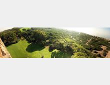 Vista panoramica dall’alto di un ampio prato verde circondato da alberi e da una fitta area boschiva, con sentieri che si diramano verso una zona più arida. Sullo sfondo si intravede la costa e il mare in lontananza.