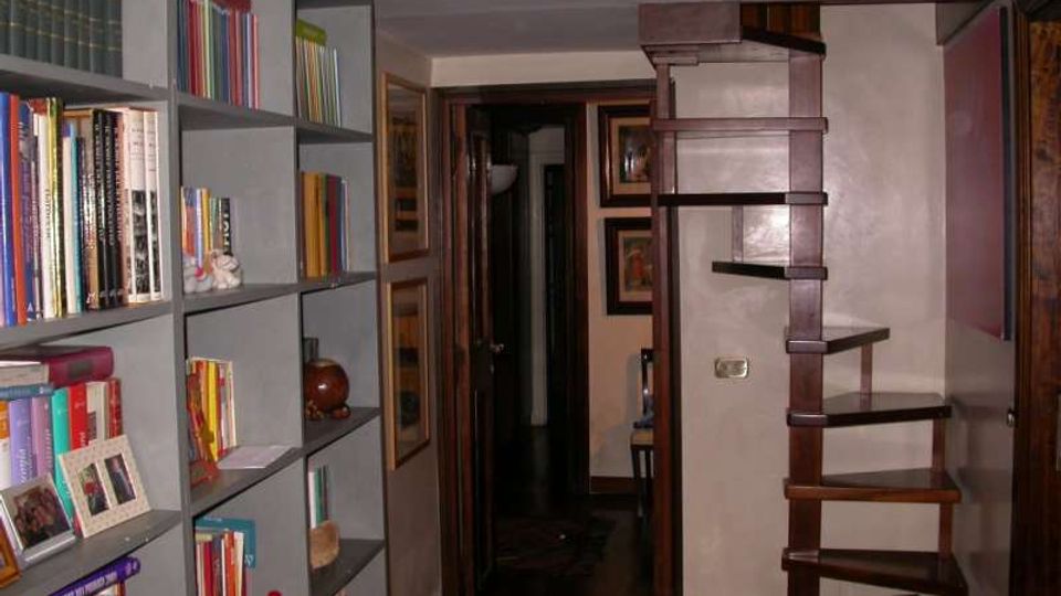 Interno di un’abitazione con corridoio stretto: a sinistra una grande libreria a scaffali pieni di libri e oggetti, a destra una scala in legno con gradini aperti. Pareti con cornici e porte in legno, pavimento scuro e soffitto basso.