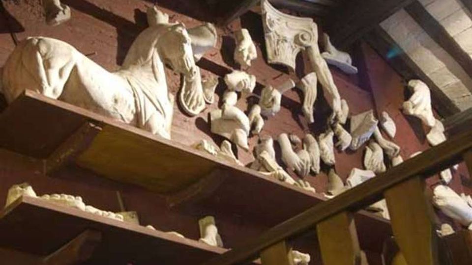 Interno di un laboratorio o magazzino con scaffalature in legno cariche di frammenti di sculture e calchi in gesso, tra cui teste e arti, e una grande testa di cavallo. Sullo sfondo si vede un soffitto con travi e pannelli, illuminato da luce artificiale.
