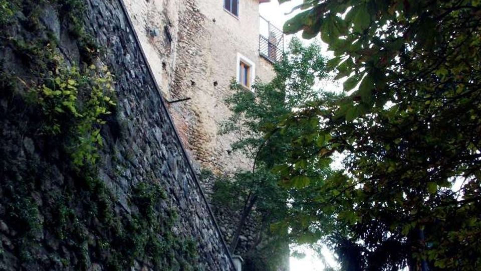 Scalinata in pietra che sale tra alti muri di contenimento in muratura, accanto a un edificio antico in pietra e intonaco con piccole finestre. Alberi e vegetazione fitta incorniciano il percorso, che conduce a una terrazza o belvedere con muretto in cima.