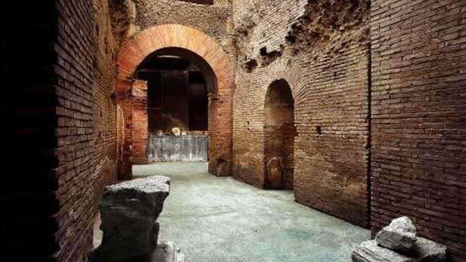 Interno di un sito archeologico con corridoio in mattoni e pietra, pareti alte con nicchie e un grande arco in laterizio che conduce a un’area più ampia. Sul pavimento in cemento si vedono blocchi di pietra e frammenti di rovine.